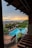 A scenic view of a resort with a large, rectangular swimming pool surrounded by lounge chairs and green palm trees. The setting includes a multi-story building with wooden railings and a sunset in the background, casting a warm glow over the ocean and distant hills.