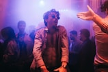 A lively nightclub scene with people dancing and socializing. The focus is on a person in sunglasses, energetically moving to the music, with several other partygoers in the background. The lighting is dim with colorful neon hues creating a vibrant atmosphere.