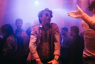 A lively nightclub scene with people dancing and socializing. The focus is on a person in sunglasses, energetically moving to the music, with several other partygoers in the background. The lighting is dim with colorful neon hues creating a vibrant atmosphere.