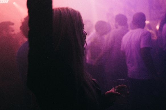 A dimly lit, crowded club scene with a woman in the foreground holding a drink and raising one arm, surrounded by people dressed casually, with a smoky, pinkish-purple hue.
