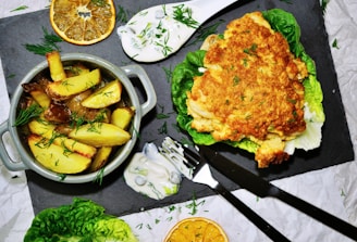 A dish features crispy potato wedges garnished with fresh dill in a small grey pot. Accompanying the potatoes is a golden-brown lasagna piece served on a bed of lettuce. There are also slices of dried orange, dollops of creamy sauce, and some scattered herbs. Cutlery is placed nearby on a dark slate serving board.