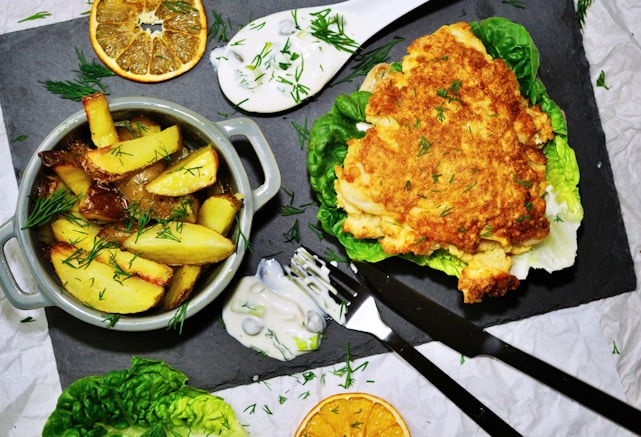 A dish features crispy potato wedges garnished with fresh dill in a small grey pot. Accompanying the potatoes is a golden-brown lasagna piece served on a bed of lettuce. There are also slices of dried orange, dollops of creamy sauce, and some scattered herbs. Cutlery is placed nearby on a dark slate serving board.