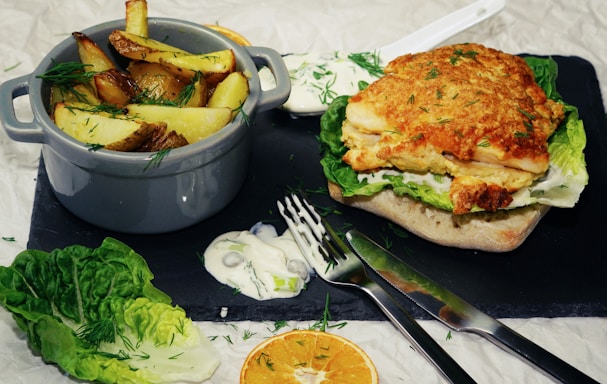 A rustic dish featuring a sandwich with crispy fried chicken on a ciabatta roll layered with fresh lettuce and creamy sauce. Alongside, a small gray pot contains golden brown potato wedges garnished with dill. There's a side of sauce spilled on a slate board, accompanied by a fork and knife. Bits of fresh dill and slices of dried orange decorate the scene, adding a touch of color to the earthy tones.