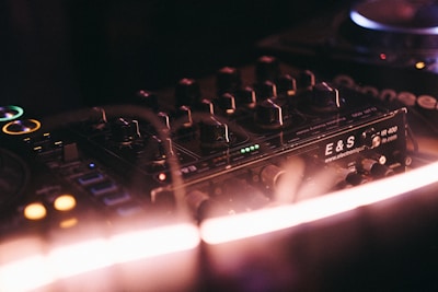 A close-up of a DJ mixer with several knobs and buttons illuminated by colorful lights. The focus is on the control panel marked with 'E&S' and other text, with soft blur highlighting the ambient atmosphere.