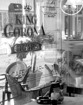A black and white image features the interior of a cafe or bar named 'King Corona Cigars.' The establishment has a retro, classic look with a person sitting inside by the window, deeply engaged in reading a newspaper while holding a cigar. Various chairs and tables are visible, along with a detailed sign on the glass panel advertising cigars. The ambiance suggests a relaxed setting.