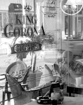 A black and white image features the interior of a cafe or bar named 'King Corona Cigars.' The establishment has a retro, classic look with a person sitting inside by the window, deeply engaged in reading a newspaper while holding a cigar. Various chairs and tables are visible, along with a detailed sign on the glass panel advertising cigars. The ambiance suggests a relaxed setting.