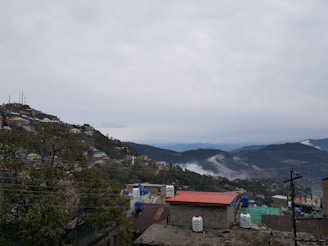 A hilly landscape featuring densely packed buildings on a slope with some construction in progress. Radio towers protrude from the top of the hill, and trees are spread across the area. In the distance, layers of rolling hills and mountains are partly covered with mist. The overcast sky adds a muted atmosphere to the scene.