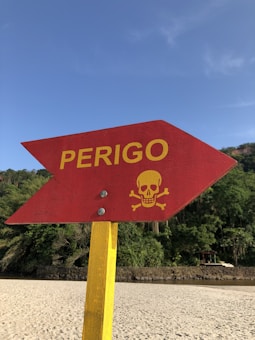 A red arrow-shaped sign with the word 'PERIGO' and a yellow skull and crossbones symbol. It is mounted on a yellow post and is situated on a sandy beach with trees and a blue sky in the background.