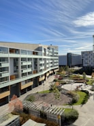 A modern residential building with multiple floors and large windows overlooks a landscaped courtyard. The area features paved pathways, greenery, and a small playground. The sky is clear with wispy clouds and there are additional modern buildings in the background.