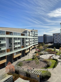 A modern residential building with multiple floors and large windows overlooks a landscaped courtyard. The area features paved pathways, greenery, and a small playground. The sky is clear with wispy clouds and there are additional modern buildings in the background.