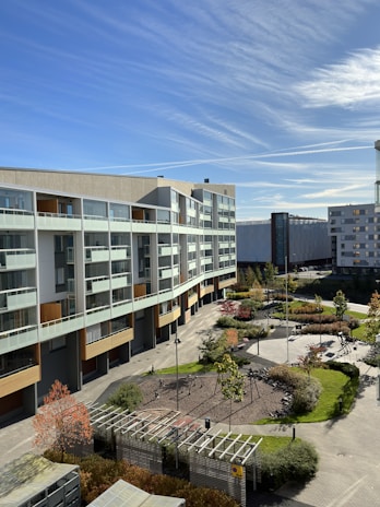 A modern residential building with multiple floors and large windows overlooks a landscaped courtyard. The area features paved pathways, greenery, and a small playground. The sky is clear with wispy clouds and there are additional modern buildings in the background.