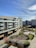 A modern residential building with multiple floors and large windows overlooks a landscaped courtyard. The area features paved pathways, greenery, and a small playground. The sky is clear with wispy clouds and there are additional modern buildings in the background.