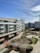 A modern residential building with multiple floors and large windows overlooks a landscaped courtyard. The area features paved pathways, greenery, and a small playground. The sky is clear with wispy clouds and there are additional modern buildings in the background.