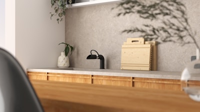 A modern kitchen interior featuring a simple design. The countertop is light-colored with a black faucet on the left side. A small potted plant in a decorative vase adds a touch of greenery. Wooden chopping boards are neatly stacked against the backsplash, complementing the wooden texture of the cabinets. The overall ambiance is minimalist and calm, with natural elements present.