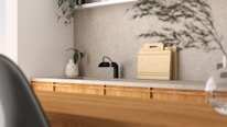 A modern kitchen interior featuring a simple design. The countertop is light-colored with a black faucet on the left side. A small potted plant in a decorative vase adds a touch of greenery. Wooden chopping boards are neatly stacked against the backsplash, complementing the wooden texture of the cabinets. The overall ambiance is minimalist and calm, with natural elements present.