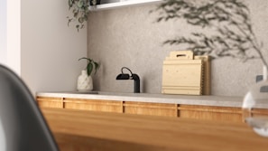 A modern kitchen interior featuring a simple design. The countertop is light-colored with a black faucet on the left side. A small potted plant in a decorative vase adds a touch of greenery. Wooden chopping boards are neatly stacked against the backsplash, complementing the wooden texture of the cabinets. The overall ambiance is minimalist and calm, with natural elements present.
