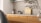 A modern kitchen interior featuring a simple design. The countertop is light-colored with a black faucet on the left side. A small potted plant in a decorative vase adds a touch of greenery. Wooden chopping boards are neatly stacked against the backsplash, complementing the wooden texture of the cabinets. The overall ambiance is minimalist and calm, with natural elements present.