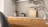 A modern kitchen interior featuring a simple design. The countertop is light-colored with a black faucet on the left side. A small potted plant in a decorative vase adds a touch of greenery. Wooden chopping boards are neatly stacked against the backsplash, complementing the wooden texture of the cabinets. The overall ambiance is minimalist and calm, with natural elements present.