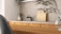 A modern kitchen interior featuring a simple design. The countertop is light-colored with a black faucet on the left side. A small potted plant in a decorative vase adds a touch of greenery. Wooden chopping boards are neatly stacked against the backsplash, complementing the wooden texture of the cabinets. The overall ambiance is minimalist and calm, with natural elements present.