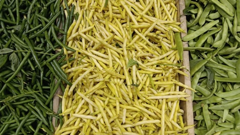 A variety of beans are displayed in a wooden crate, with yellow beans in the center, surrounded by green beans on both sides. The vibrant colors and textures of the fresh produce are clearly visible.