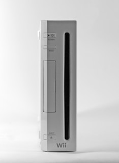 A white video game console stands vertically against a plain background. The console features buttons labeled power, reset, and eject, with a narrow slot indicating where discs are inserted. The design is sleek and minimalistic.