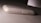 A white or light-colored smartphone is lying face down on a textured surface. The focus is on the back cover and side edges of the phone, highlighting the USB port and a small hole, possibly for a microphone or speaker. The surface beneath the phone looks slightly dirty, and the phone itself shows signs of wear.