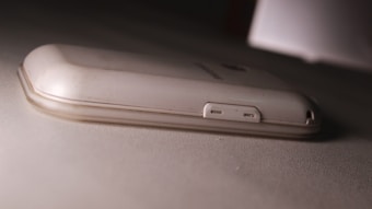 A white or light-colored smartphone is lying face down on a textured surface. The focus is on the back cover and side edges of the phone, highlighting the USB port and a small hole, possibly for a microphone or speaker. The surface beneath the phone looks slightly dirty, and the phone itself shows signs of wear.