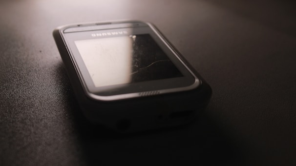 A vintage smartphone is lying on a dark surface, with a slightly reflective screen and a visible brand name above the display. The phone's surface has scuff marks, indicating wear and tear.