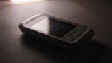 A vintage Samsung mobile phone with physical buttons is placed on a smooth, dark surface. The lighting is dim, creating a nostalgic and subdued atmosphere, casting soft shadows around the device.