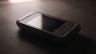 A vintage Samsung mobile phone with physical buttons is placed on a smooth, dark surface. The lighting is dim, creating a nostalgic and subdued atmosphere, casting soft shadows around the device.