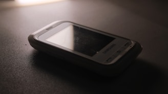 A vintage Samsung mobile phone with physical buttons is placed on a smooth, dark surface. The lighting is dim, creating a nostalgic and subdued atmosphere, casting soft shadows around the device.