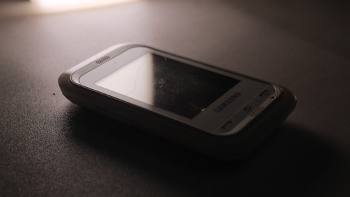 A vintage Samsung mobile phone with physical buttons is placed on a smooth, dark surface. The lighting is dim, creating a nostalgic and subdued atmosphere, casting soft shadows around the device.