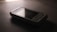 A vintage Samsung mobile phone with physical buttons is placed on a smooth, dark surface. The lighting is dim, creating a nostalgic and subdued atmosphere, casting soft shadows around the device.