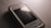 A close-up of an older Samsung mobile phone with a scratched screen placed on a textured surface. The phone's case is slightly transparent, and there is a reflection of light on the screen.