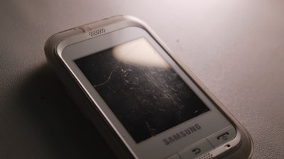 A close-up of an older Samsung mobile phone with a scratched screen placed on a textured surface. The phone's case is slightly transparent, and there is a reflection of light on the screen.