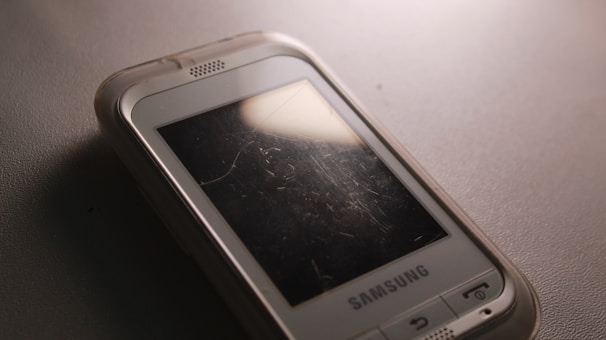 A close-up of an older Samsung mobile phone with a scratched screen placed on a textured surface. The phone's case is slightly transparent, and there is a reflection of light on the screen.
