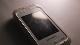 A close-up of an older Samsung mobile phone with a scratched screen placed on a textured surface. The phone's case is slightly transparent, and there is a reflection of light on the screen.