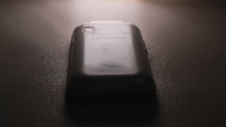 A slightly worn smartphone with a visible Samsung logo on the back, placed on a flat, dark surface under low lighting conditions. The light source is coming from the top, casting subtle shadows around the phone.