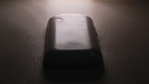 A slightly worn smartphone with a visible Samsung logo on the back, placed on a flat, dark surface under low lighting conditions. The light source is coming from the top, casting subtle shadows around the phone.