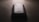 A slightly worn smartphone with a visible Samsung logo on the back, placed on a flat, dark surface under low lighting conditions. The light source is coming from the top, casting subtle shadows around the phone.