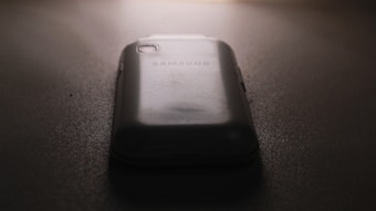 A slightly worn smartphone with a visible Samsung logo on the back, placed on a flat, dark surface under low lighting conditions. The light source is coming from the top, casting subtle shadows around the phone.