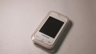 A small, older model Samsung smartphone with a white exterior and a black screen lies on a flat, light gray surface. The device has a physical home button and a compact design.