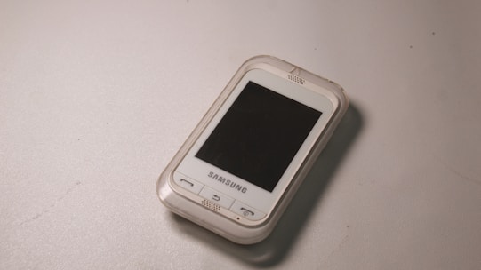 A small, older model Samsung smartphone with a white exterior and a black screen lies on a flat, light gray surface. The device has a physical home button and a compact design.
