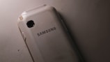 A slightly worn Samsung smartphone lying on a textured surface. The phone is positioned with its back facing up, displaying the brand name prominently in the center. The surface and edges of the phone show some signs of wear and scratches.