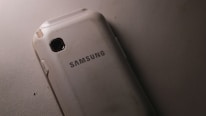 A slightly worn Samsung smartphone lying on a textured surface. The phone is positioned with its back facing up, displaying the brand name prominently in the center. The surface and edges of the phone show some signs of wear and scratches.