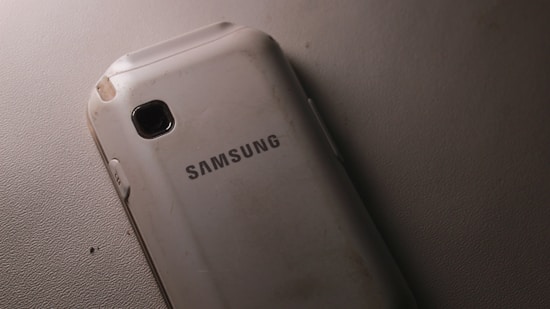 A slightly worn Samsung smartphone lying on a textured surface. The phone is positioned with its back facing up, displaying the brand name prominently in the center. The surface and edges of the phone show some signs of wear and scratches.