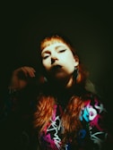 A person with red hair and dramatic makeup is bathed in shadow, with a beam of light illuminating part of their face. They wear a colorful patterned outfit, featuring bright colors like purple, blue, and pink. The expression is intense, with bold eyeliner and dark lipstick, creating a striking contrast.