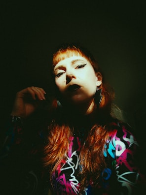 A person with red hair and dramatic makeup is bathed in shadow, with a beam of light illuminating part of their face. They wear a colorful patterned outfit, featuring bright colors like purple, blue, and pink. The expression is intense, with bold eyeliner and dark lipstick, creating a striking contrast.