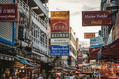A bustling urban street filled with various signs in multiple languages, indicating different businesses such as restaurants and car parks. The scene is characterized by a mix of cultural influences and is crowded with overhead wires and tightly packed buildings.