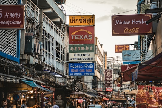 A bustling urban street filled with various signs in multiple languages, indicating different businesses such as restaurants and car parks. The scene is characterized by a mix of cultural influences and is crowded with overhead wires and tightly packed buildings.